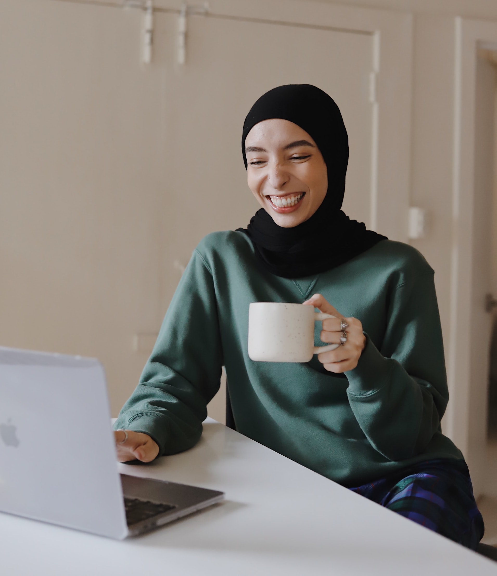 Woman with hijab sitting at a table, holding a mug and smiling, looking at laptop screen